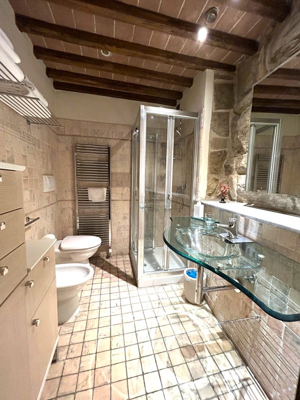 bagno-residence-epoca-arezzo Vista completa del bagno con doccia, lavabo in vetro, sanitari e soffitto con travi in legno